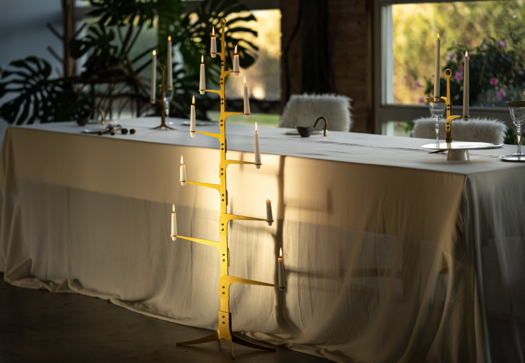 Dinertafel met oversized tafelkleed voor een raam met een plant, the ore glow serie kandelaren in sunnygold uitgevoerd staan op en voor tafel met brandende kaarsen.
